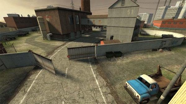 A screenshot from Counter-Strike: Source showing an industrial map with buildings, a rusty truck, and open gates.