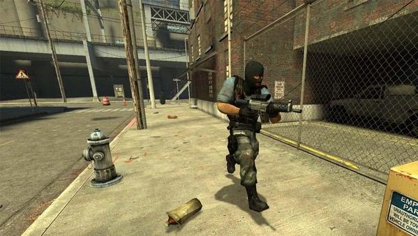 Counter-Strike: Source screenshot showing an armed player in tactical gear running on a city street near a chain-link fence.