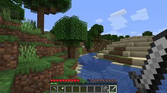Minecraft: Java Edition screenshot of a player holding a sword by a river, with grassy hills, trees, and a sandy shore in view.