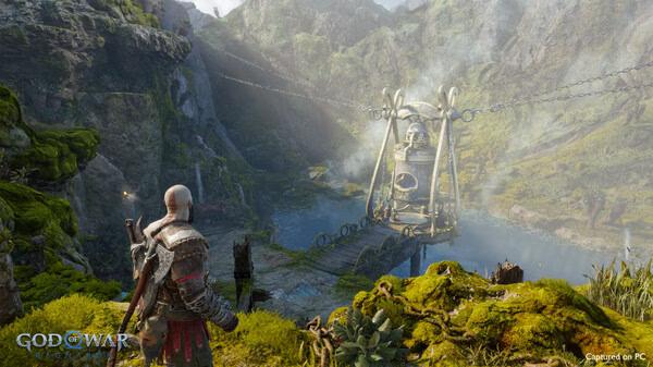 God of War Ragnarök screenshot showing Kratos standing on a green cliff, overlooking a large chained mechanism in a scenic valley.