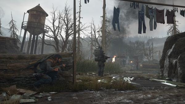 Screenshot from Days Gone: A character crouches with a rifle near a water tower, while another walks ahead past hanging laundry in a desolate landscape.
