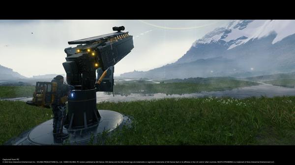 Screenshot from DEATH STRANDING DIRECTOR'S CUT showing a character standing beside a large cargo-launching device in a grassy landscape.