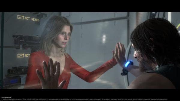 Screenshot from DEATH STRANDING DIRECTOR'S CUT shows two characters touching hands across a glass partition in a futuristic setting.