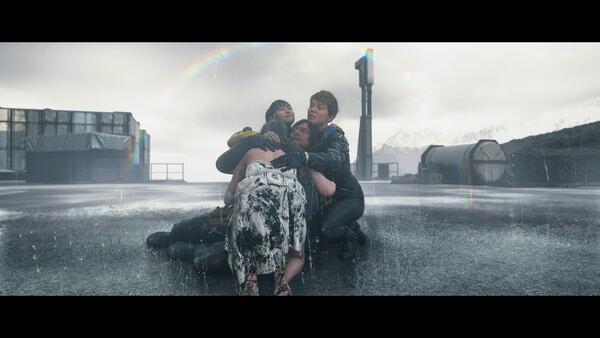 Screenshot from DEATH STRANDING 2: ON THE BEACH showing a group of characters huddled together in the rain on a desolate landscape.