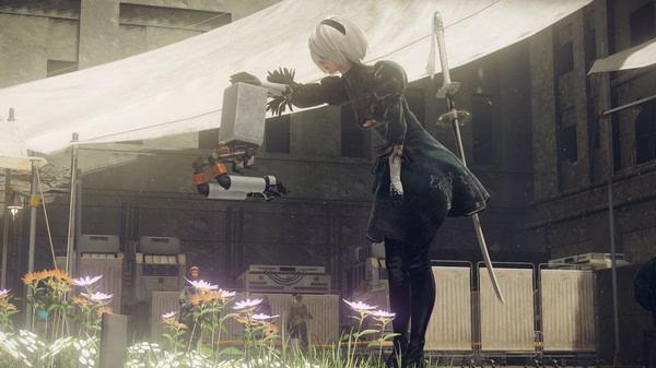 Screenshot from NieR:Automata™ showing a character interacting with a robot in a garden with flowers and tents visible.