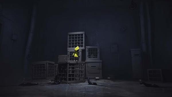 Little Nightmares screenshot: A small figure in a yellow raincoat stands on a stack of cages in a dimly lit, eerie room.