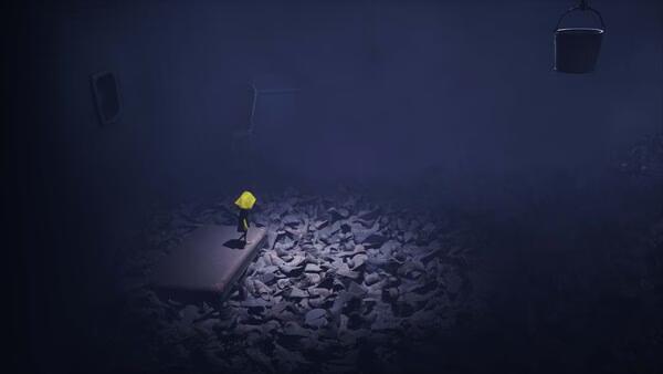 Screenshot from Little Nightmares showing a small figure in a yellow raincoat standing on a mattress amidst scattered shoes in a dark room.