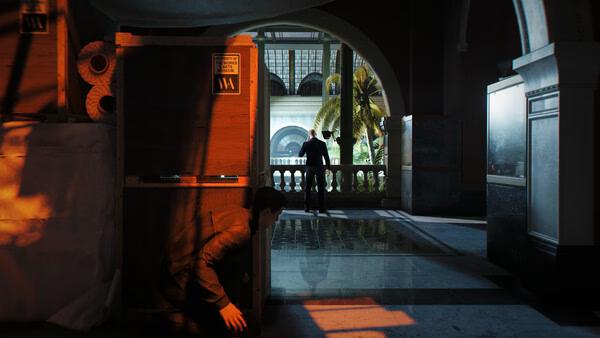 007 First Light screenshot: A character crouches behind crates in a dimly lit area, observing another figure on a balcony.