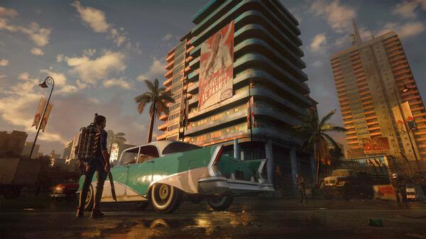 A Far Cry® 6 screenshot showing a character near a vintage car in a city square with tall buildings and a political banner.