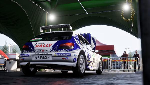 Screenshot from Assetto Corsa Rally: A rally car is parked under a green canopy in a service area.