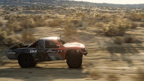 BeamNG.drive screenshot showing a racing truck with graphic decals speeding through a desert landscape, surrounded by shrubs.