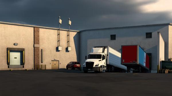 American Truck Simulator screenshot: A white semi-truck with a trailer parked near a warehouse loading dock under a cloudy sky.