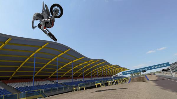 MX Bikes screenshot: A motocross rider performs a high jump stunt in a stadium with empty stands under a clear blue sky.
