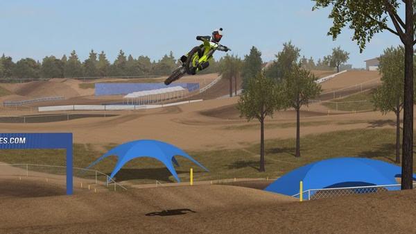 MX Bikes screenshot: A motorcyclist in mid-air performs a jump over a dirt track, surrounded by trees and blue tents.