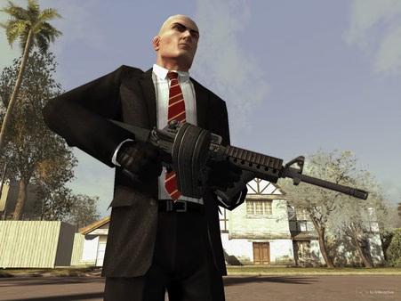 Hitman: Blood Money screenshot of Agent 47 holding a large weapon outdoors, wearing a suit and gloves, with houses in the background.