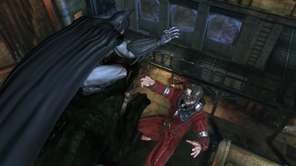 Batman: Arkham Asylum screenshot showing Batman lunging at an enemy in red robes within a dimly lit industrial setting.