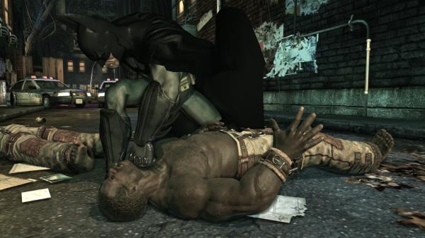 Batman: Arkham Asylum screenshot showing Batman restraining a man on the ground in a gritty, dimly lit alleyway with police cars nearby.