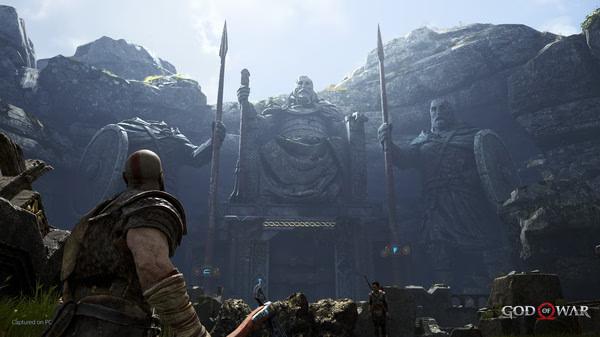 Kratos stands before a towering statue of Norse gods in a rocky landscape in this 'God of War' screenshot.