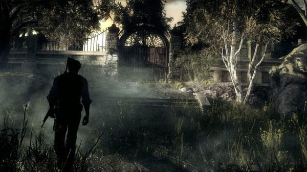 A lone figure walks through a foggy, eerie forest in this screenshot from "The Evil Within", with trees and a gated entrance ahead.