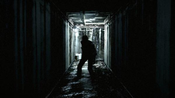 A screenshot from The Evil Within shows a silhouetted figure standing in a dimly lit, eerie hallway with water dripping from the ceiling.