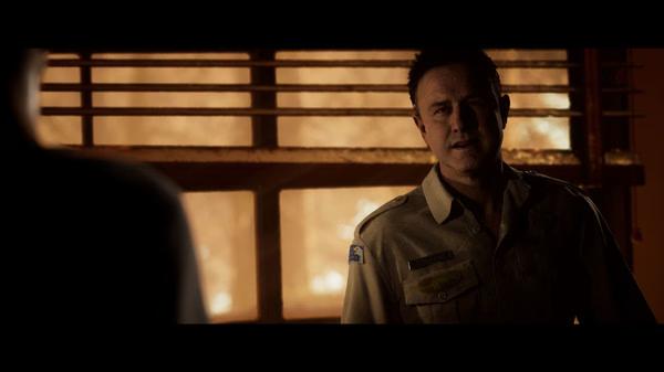 A screenshot from The Quarry showing a character in a sheriff's uniform standing near a window with warm lighting.