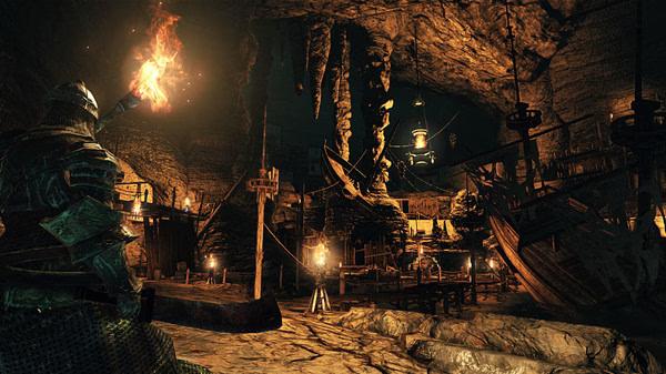 Dark Souls II: Scholar of the First Sin screenshot showing a knight holding a torch in a dimly lit, cavernous area with hanging boats.