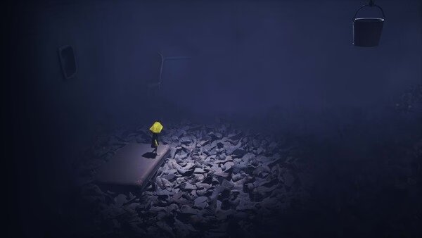 Screenshot from Little Nightmares showing a small figure in a yellow raincoat standing on a mattress amidst scattered shoes in a dark room.