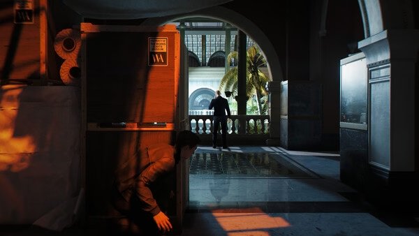 007 First Light screenshot: A character crouches behind crates in a dimly lit area, observing another figure on a balcony.