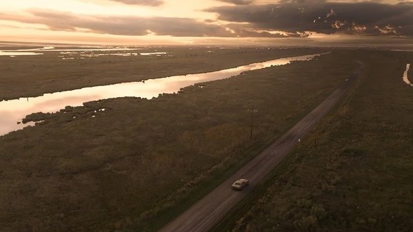A screenshot from Resident Evil 7 Biohazard showing a car driving down a rural road at sunset near a river.