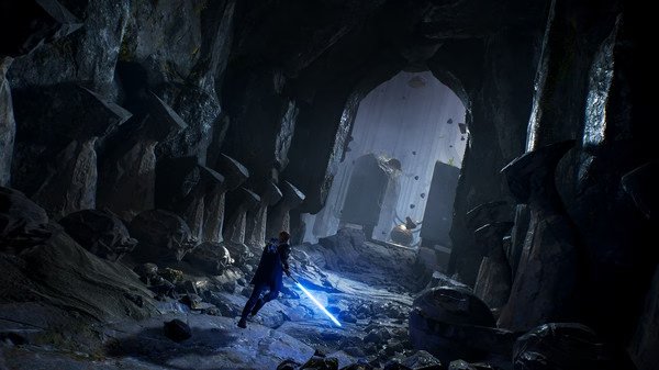 A screenshot from STAR WARS Jedi: Fallen Order showing the protagonist wielding a blue lightsaber in a dark, rocky cavern.