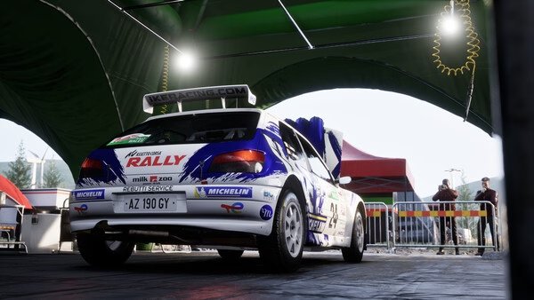 Screenshot from Assetto Corsa Rally: A rally car is parked under a green canopy in a service area.