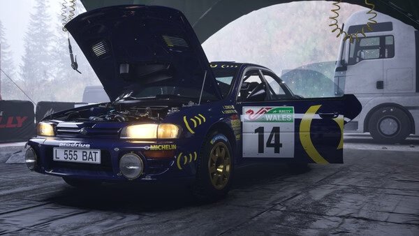 A screenshot from Assetto Corsa Rally shows a blue rally car with its hood open in a garage, marked with the number 14.