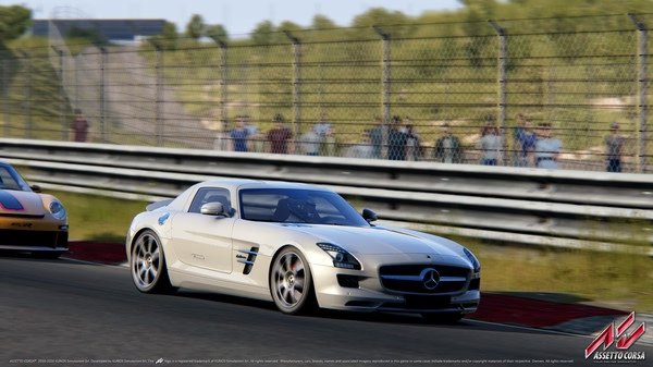 Assetto Corsa screenshot: A white sports car races down a sunny track, with spectators behind a fence watching intently.