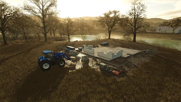 Farming Simulator 25 screenshot: A blue tractor beside a partially built structure on a farm, surrounded by trees and a river.
