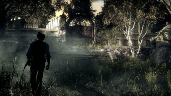 A lone figure walks through a foggy, eerie forest in this screenshot from "The Evil Within", with trees and a gated entrance ahead.