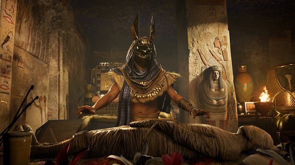 Screenshot from Assassin's Creed Origins showing a masked figure resembling Anubis with a mummy in an ancient Egyptian tomb.