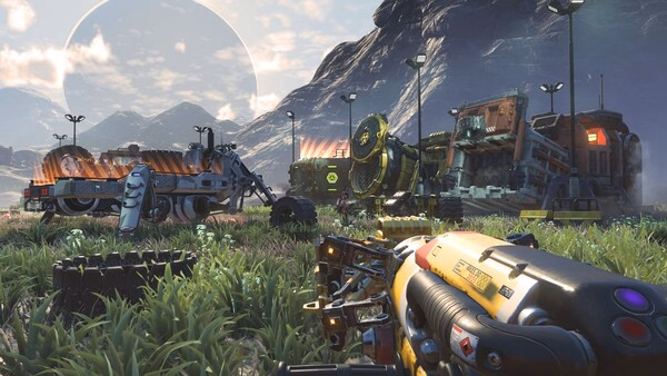 Screenshot from No Man's Sky showing a futuristic base with vehicles and machinery against a mountainous landscape.