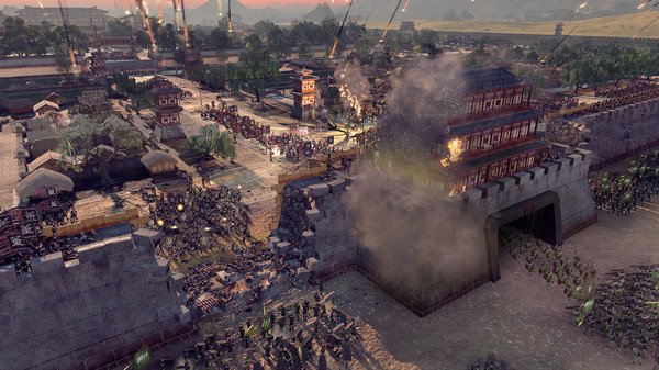 Screenshot from Total War: Three Kingdoms showing a dramatic siege with troops attacking a fortified city under heavy fire.