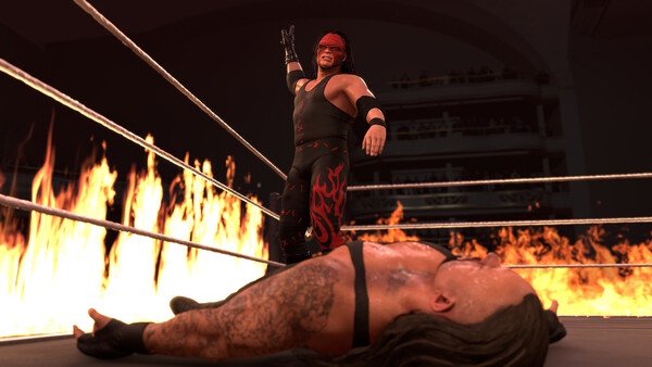 WWE 2K26 screenshot: A wrestler in a black and red outfit stands triumphantly over a fallen opponent in a ring surrounded by flames.