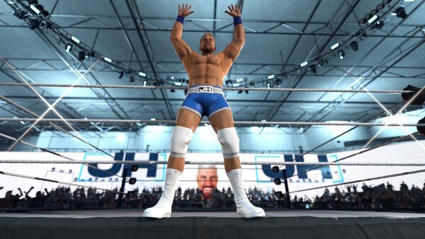 WWE 2K26 screenshot: Wrestler in blue trunks and white boots stands triumphantly in the ring, arms raised, with a cheering crowd behind.