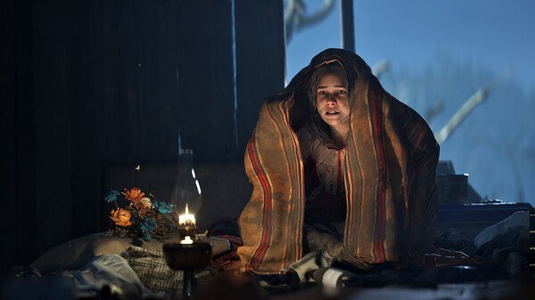 A screenshot from 'Hell is Us' showing a person wrapped in blankets near a glowing lantern, surrounded by objects in a dimly lit room.