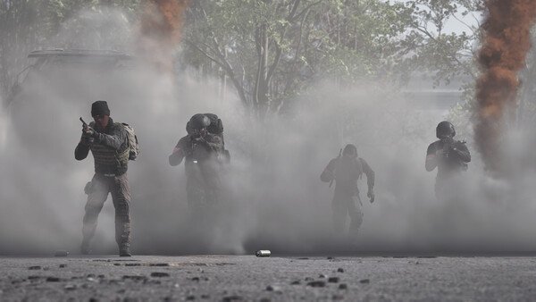 WARDOGS screenshot: Four armed soldiers emerge from dense smoke on a battlefield, with orange plumes rising in the background.